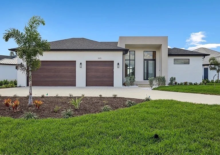 Single-story white stucco home with two wood-tone garage doors, a tall glass entry and a young palm in the front yard